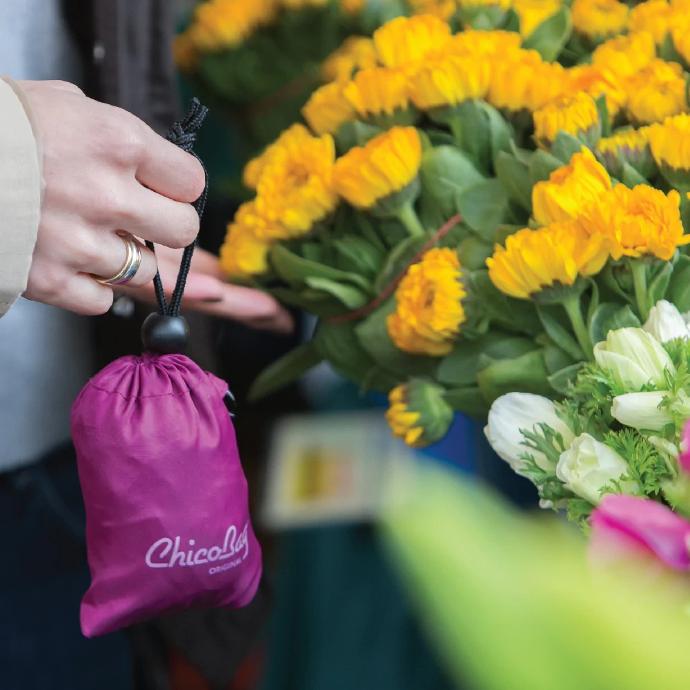Boysenberry Original reusable tote pouched hanging off of someones finger at a farmers market next to some flowers