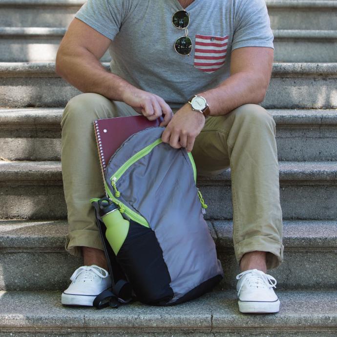 person sitting on steps placing a notebook in a ChicoBag Travel Pack rePETE pouchable backpack with a reusable water bottle sitting in the side bottle pocket