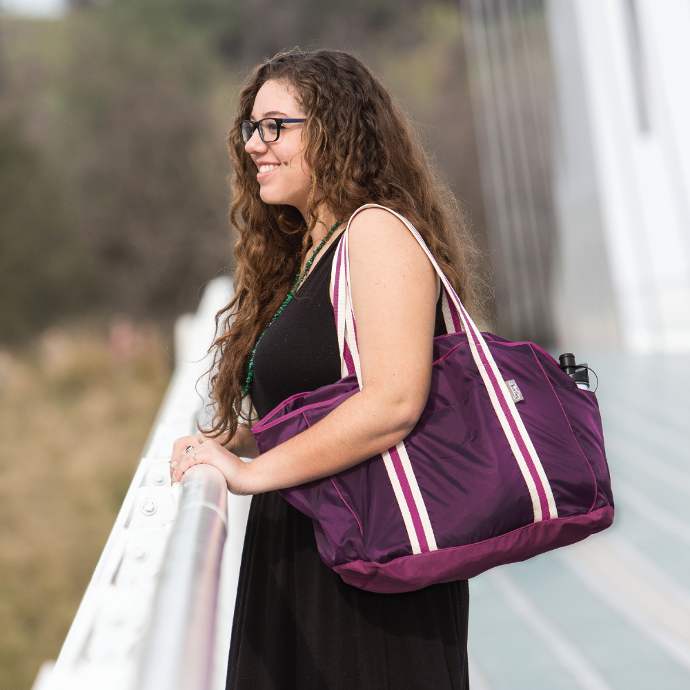 person on a bridge wearing a ChicoBag Purple Nomad packable large capacity reusable tote on their shoulder looking out into the water